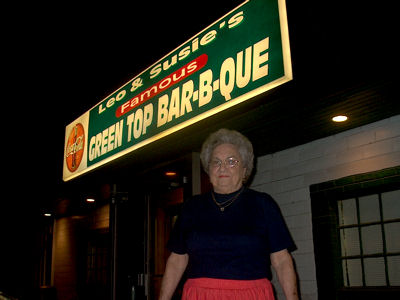Susie Headrick standing under the Leo & Susie's Famous Green Top Bar-B-Que sign at night